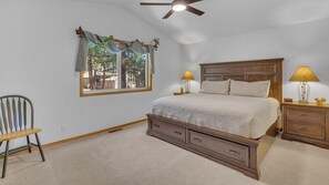 A bedroom with a large bed, two bedside tables with lamps, a chair, a window with a view of trees, and a ceiling fan. - Coyote Lane 15 (Bend)