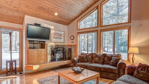 A cozy living room with a fireplace, wooden flooring, and large windows offering a view of the outdoors. - Ashwood Lane 1 (Bend)