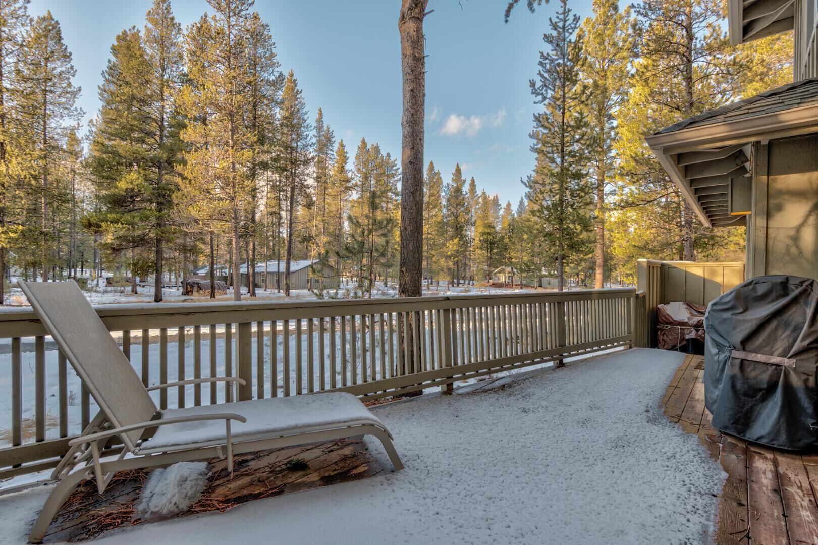 A snow-covered deck with a chair and a grill, surrounded by tall pine trees.