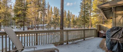 A snow-covered deck with a chair and a grill, surrounded by tall pine trees.