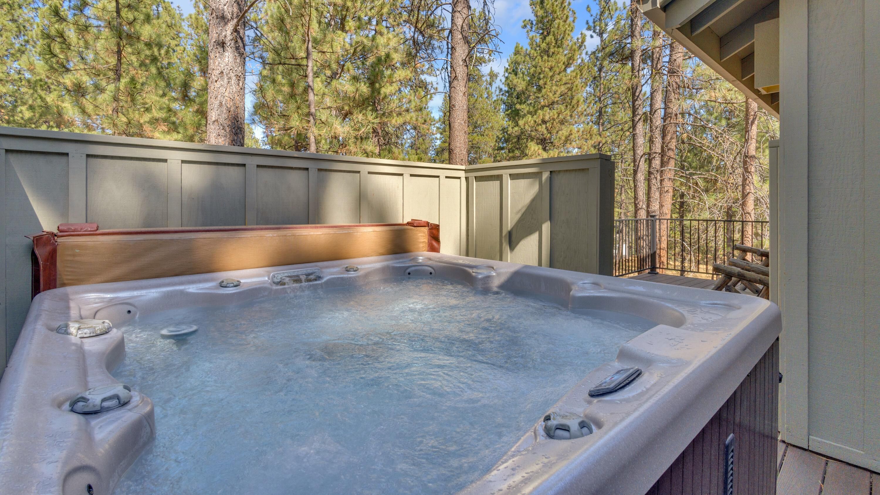 A hot tub on an outdoor deck with a wooden railing and a view of pine trees.