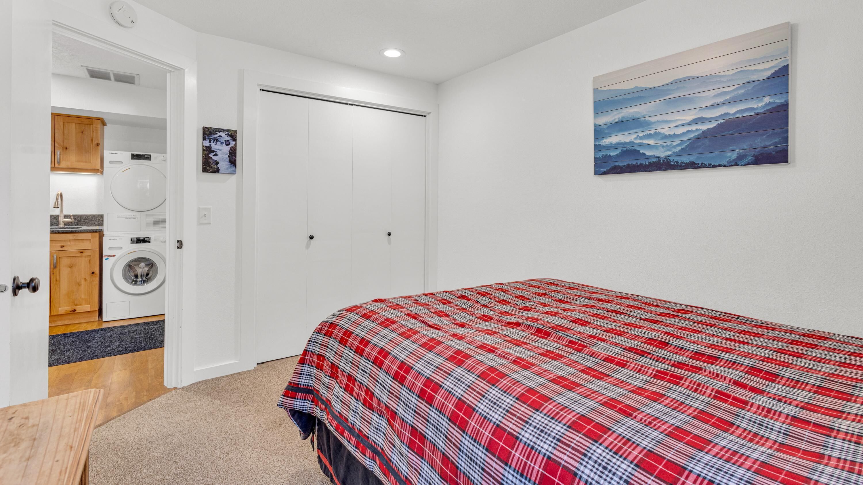A bedroom with a red plaid bedspread, a washing machine, and a painting on the wall.