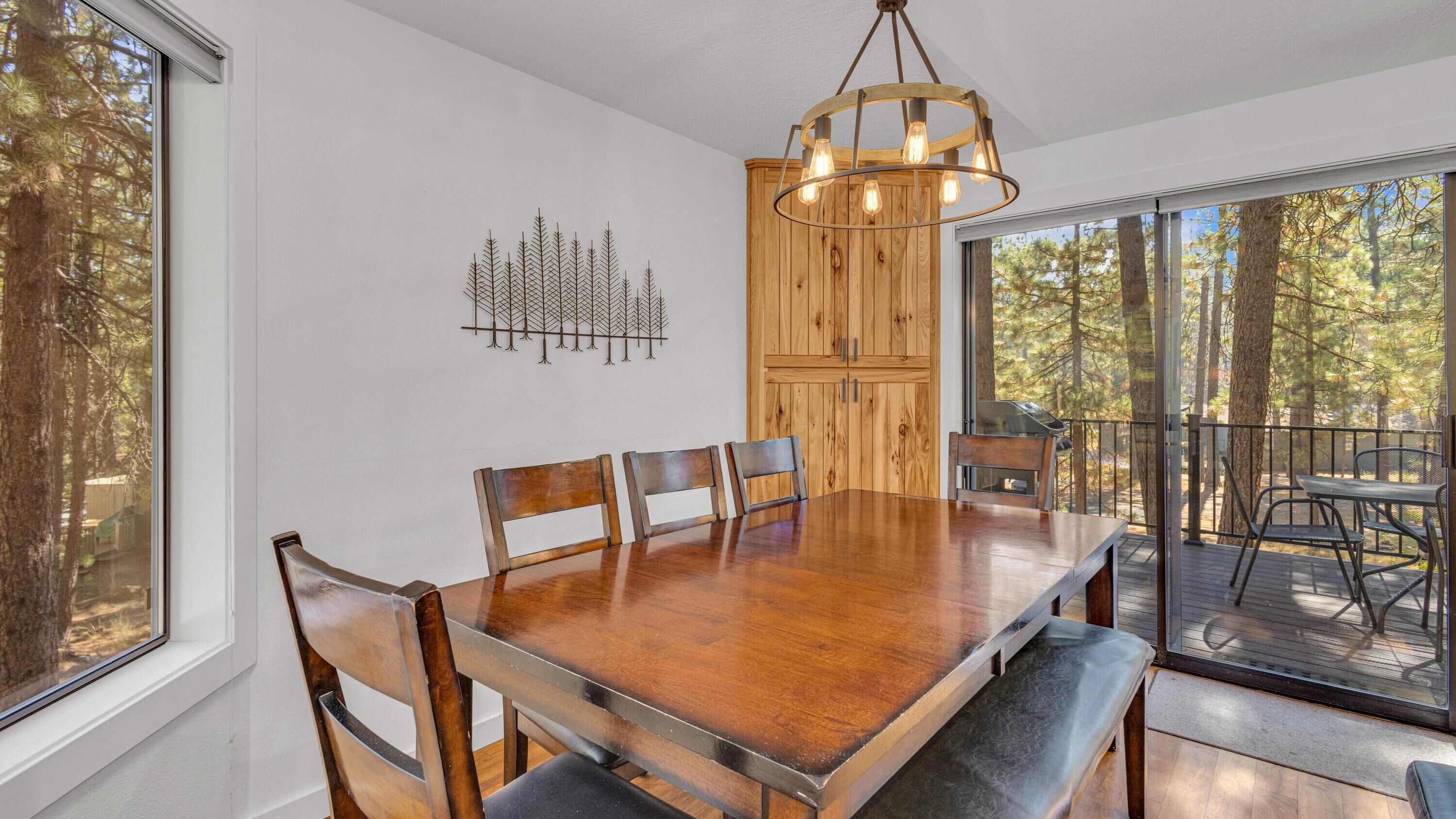 A dining area with a wooden table and chairs, a chandelier, and a view of trees through large windows.