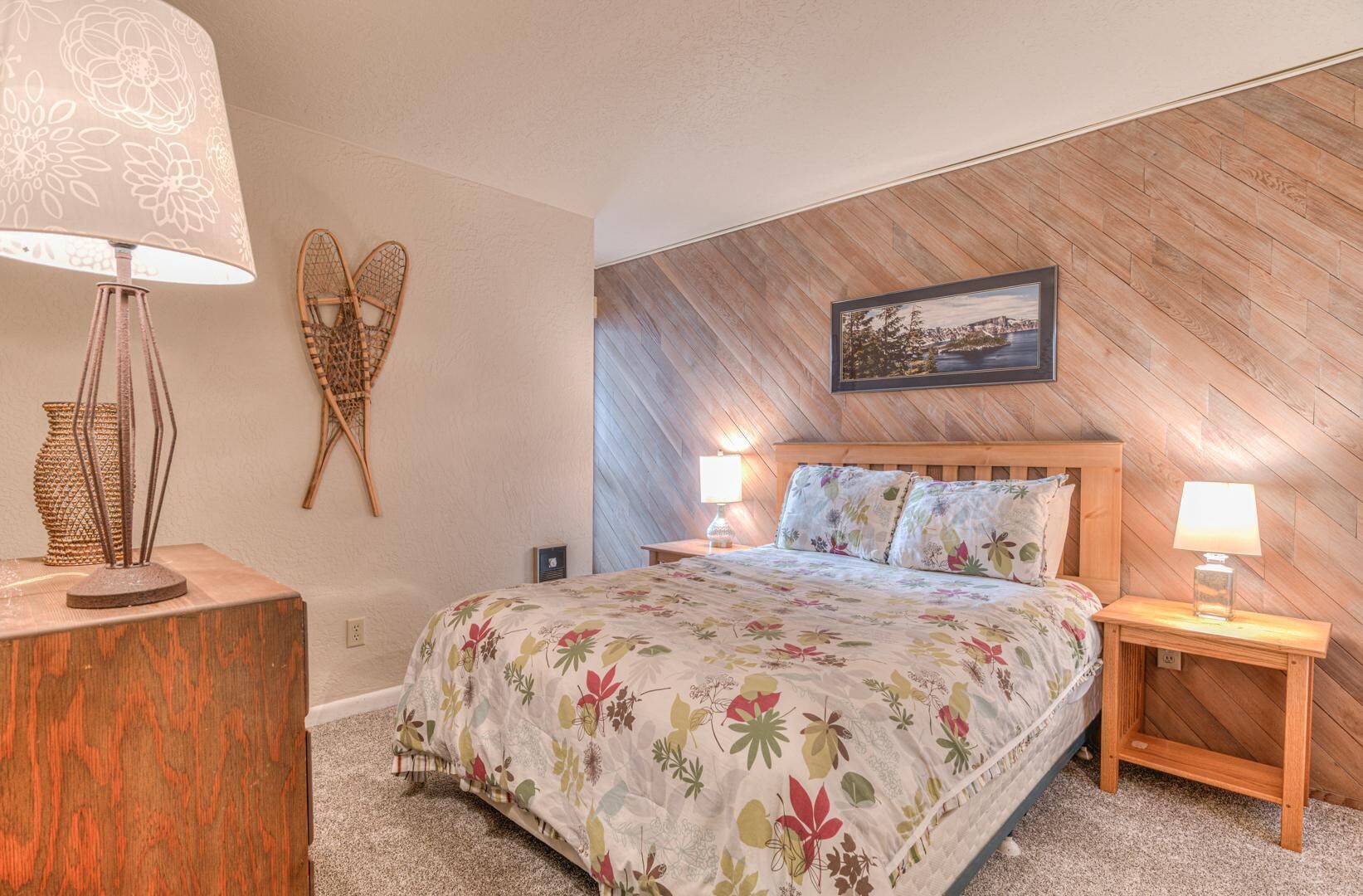 A bedroom with a wooden sloped ceiling, a bed with floral bedding, two bedside lamps, and a framed picture on the wall.