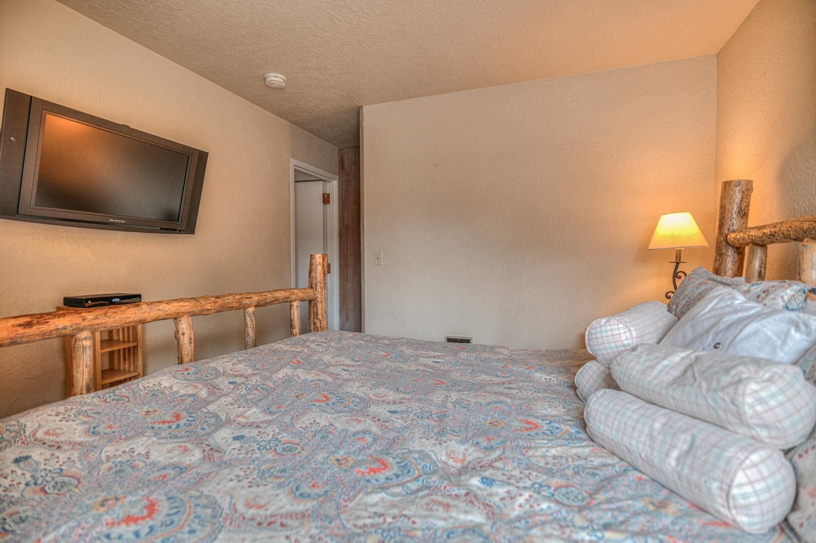 A bedroom with a bed, a television mounted on the wall, a wooden headboard, and a bedside lamp.