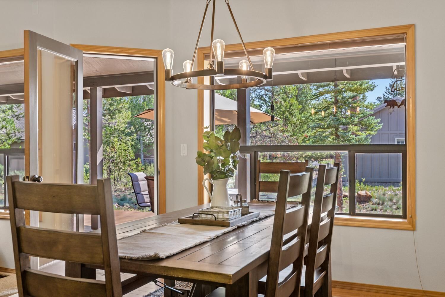 Un comedor con una mesa y sillas de madera, una araña y ventanas amplias con vistas al exterior.