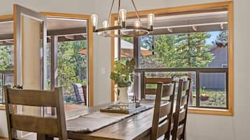Un comedor con una mesa y sillas de madera, una araña y ventanas amplias con vistas al exterior.