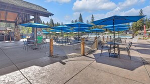 An outdoor patio area with tables and chairs under blue umbrellas, a pool, and a wooden building structure. - Lassen Lane 3 (Bend)