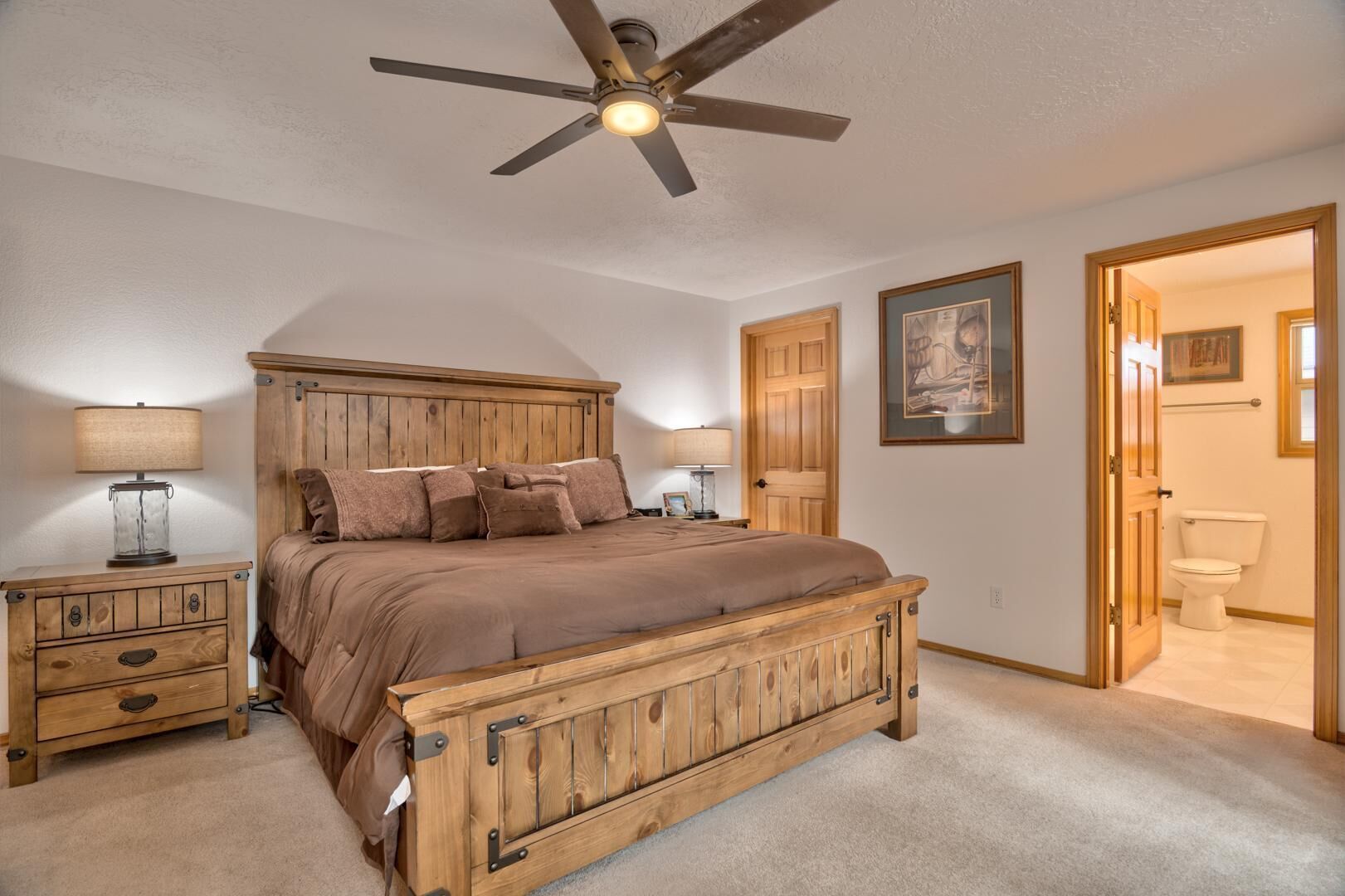 A bedroom with a wooden bed, two bedside tables, a ceiling fan, and a door leading to a bathroom.