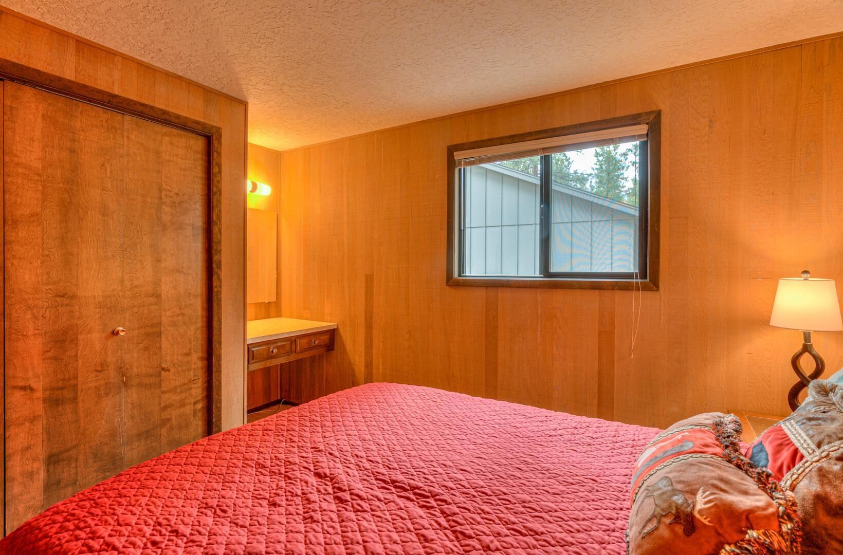 Un dormitorio con paredes de madera, una ventana, una cama con colcha roja, una mesita de noche con lámpara y un aparador con espejo.