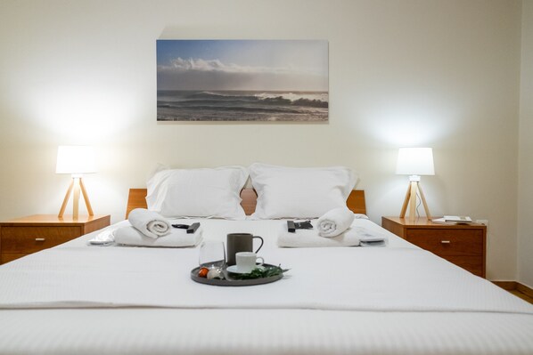 A neatly made bed with white linens, two bedside tables with lamps, a tray with a cup and a mug, and a painting of the ocean on the wall.