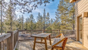 A wooden deck with a table and chairs, surrounded by a railing, overlooking a forested area.