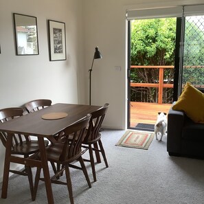 Un coin repas avec une table et des chaises en bois, un lampadaire et une porte-fenêtre coulissante donnant sur un balcon doté d’une rambarde en bois et où se trouve un chien.