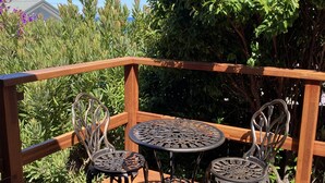 Une terrasse en bois avec une table et des chaises en métal, entourée d’une végétation luxuriante et sous un ciel bleu dégagé.