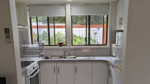 Une cuisine compacte avec des meubles blancs, un four encastré, un évier et une fenêtre donnant sur une clôture et de la verdure.