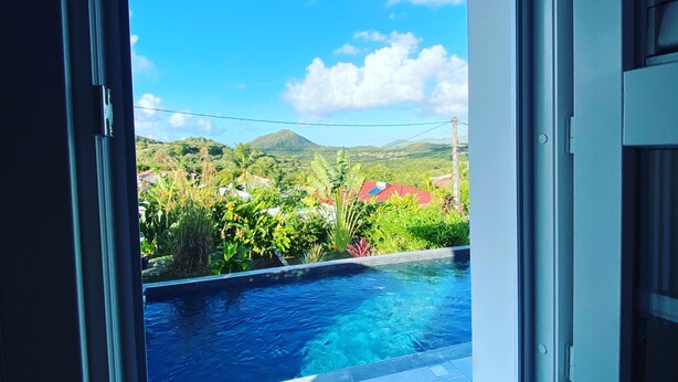 A window with a view of a swimming pool, lush greenery, and distant hills.