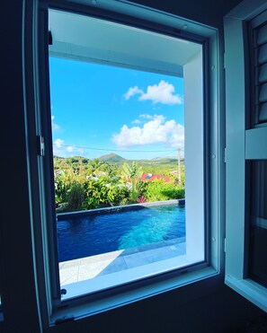 A window with a view of a swimming pool, lush greenery, and distant hills. - Comfort & privacy - 2 master suites (Sainte-Anne)
