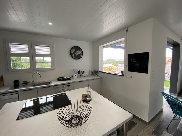 A modern kitchen with white cabinetry, a large island, a world map clock, and a window with a view of greenery. - Comfort & privacy - 2 master suites (Sainte-Anne)