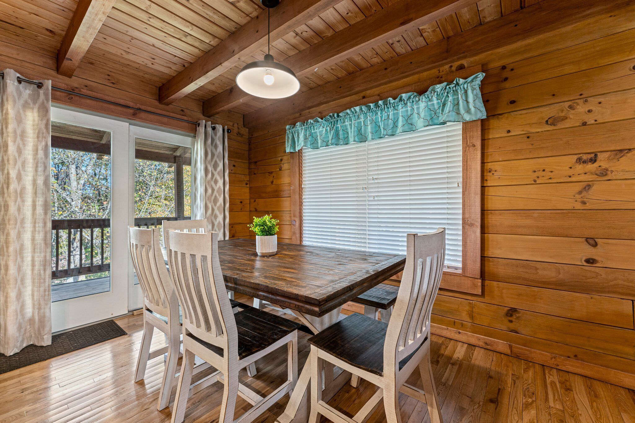 Interior de una cabaña de madera con una mesa de comedor, cuatro sillas, un ventilador de techo y una ventana con persianas.