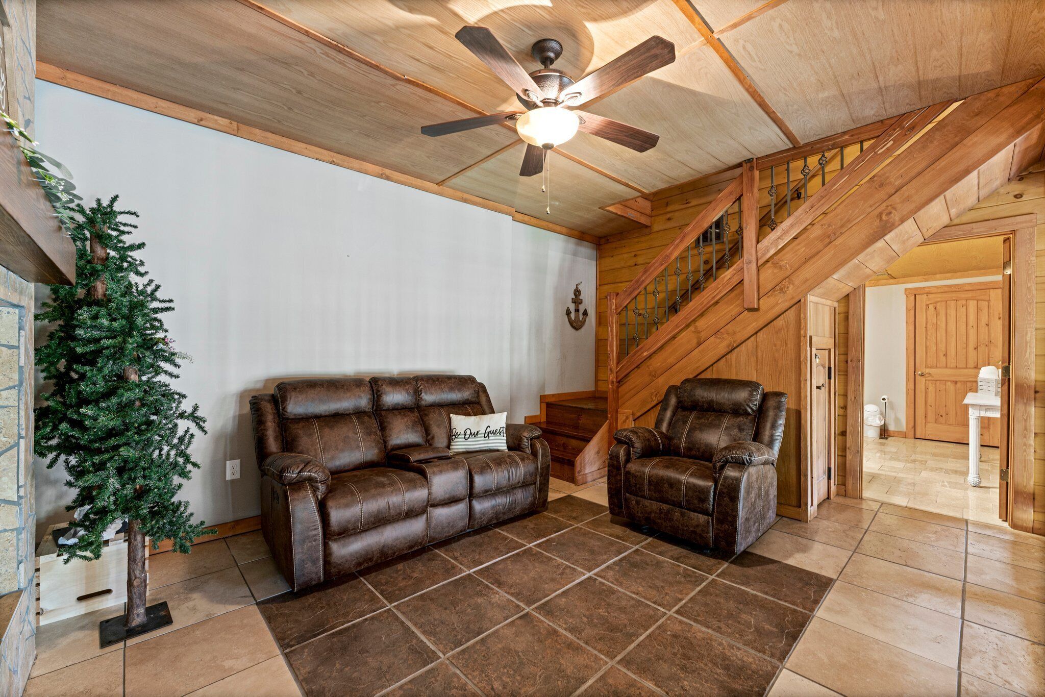 A spacious living room with a brown leather sofa, a smaller armchair, a wooden staircase, and a ceiling fan.