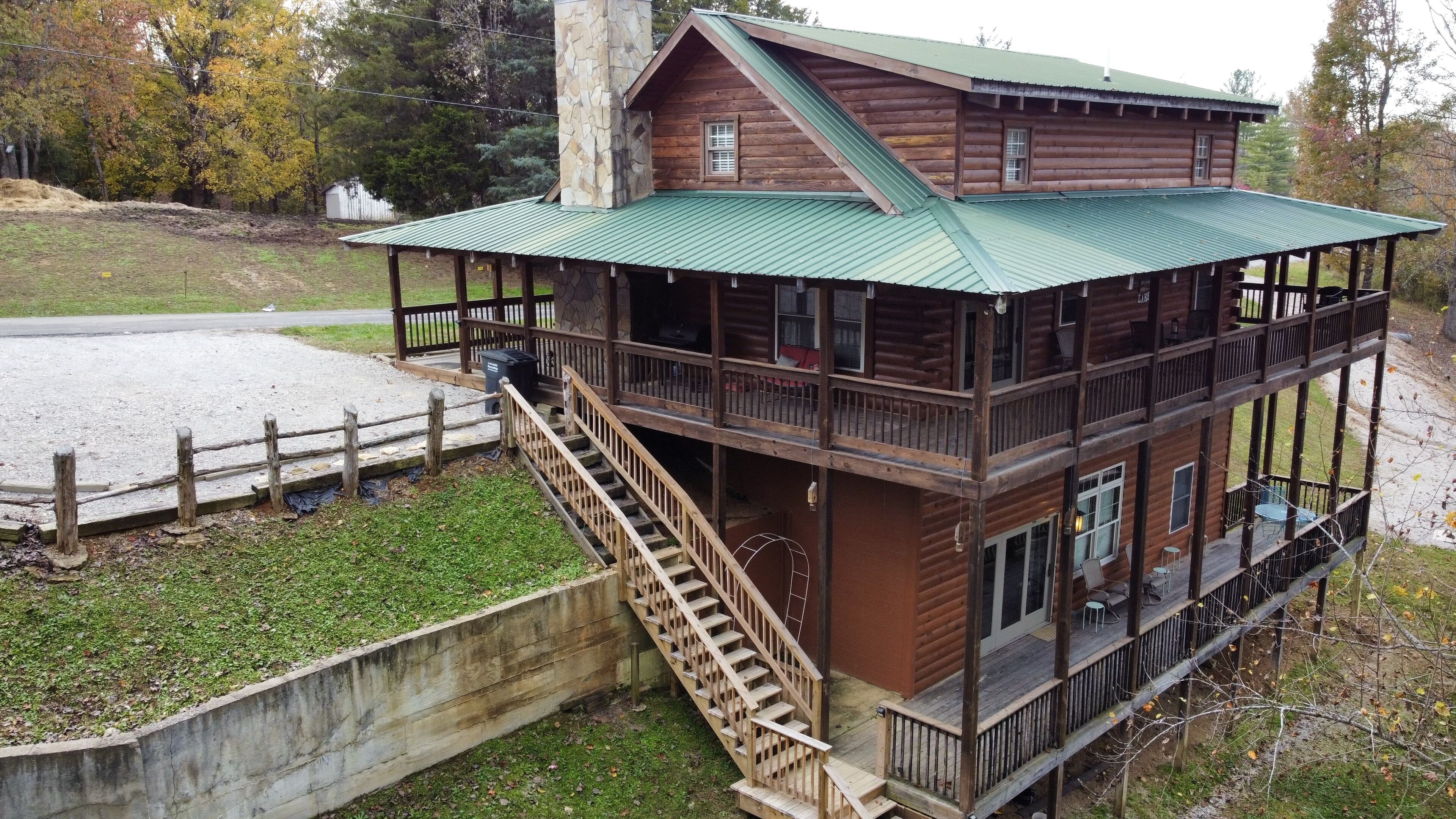 A multi-level log cabin with a green metal roof, a stone chimney, and a wooden staircase leading to the upper level.