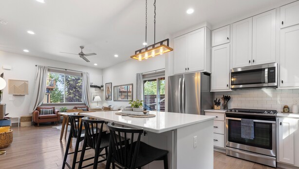 A modern kitchen with white cabinets, a central island with bar stools, stainless steel appliances, and a large window with curtains. - Bozeman/4Corners HomeBase • River Access & Fire Pit • Close to Skiing and Hiking (Bozeman)
