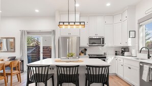 A modern kitchen with white cabinets, a central island with bar stools, stainless steel appliances, and a hanging light fixture. - Bozeman/4Corners HomeBase • River Access & Fire Pit • Close to Skiing and Hiking (Bozeman)