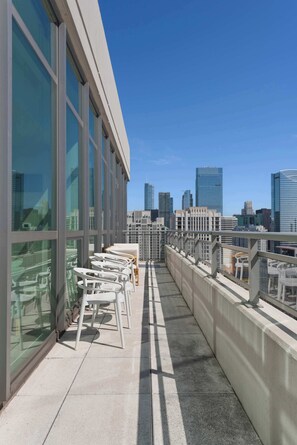 A rooftop terrace with white chairs and tables, offering a city skyline view.