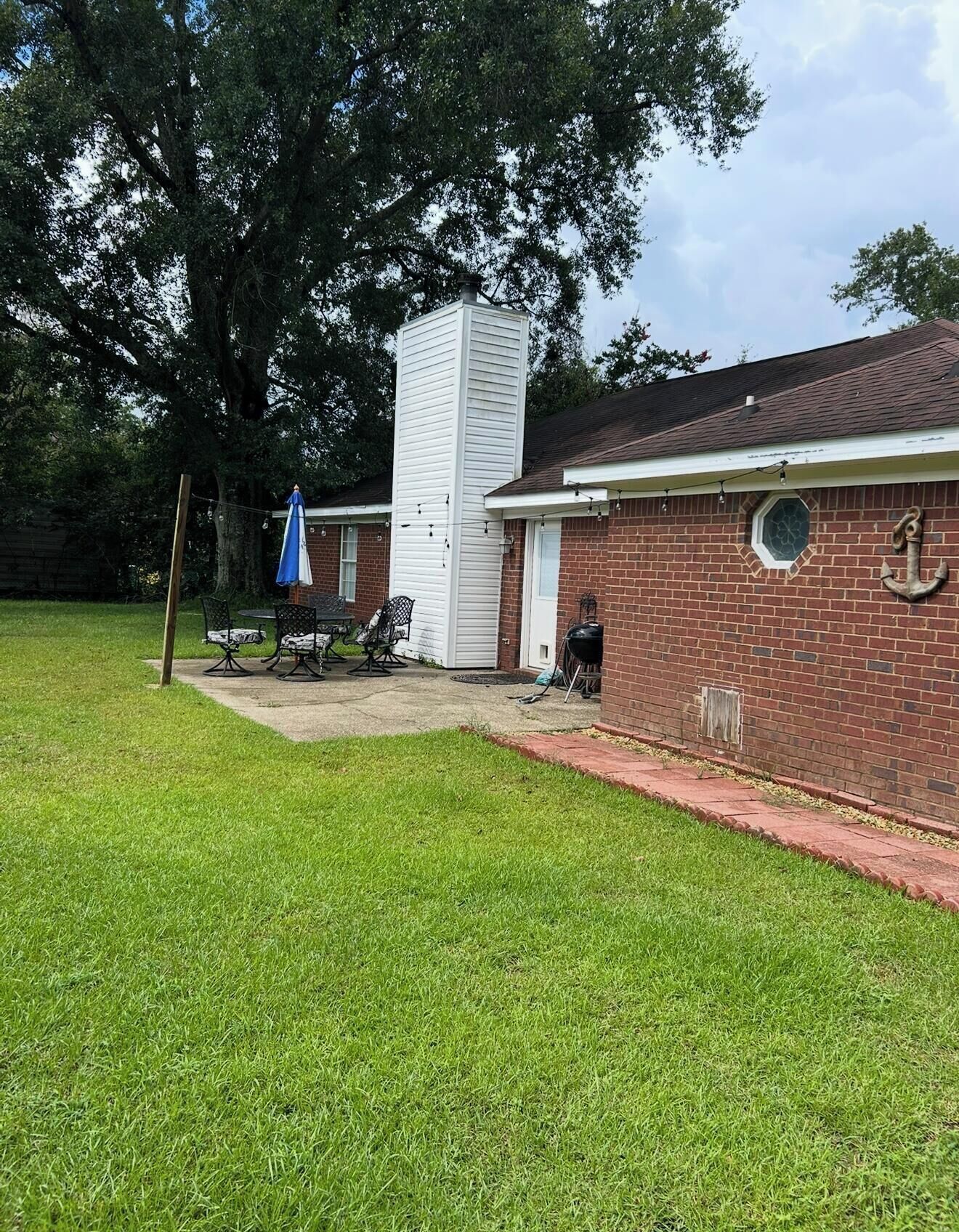 A brick house with a chimney, a covered patio with chairs, and a large tree in the background.