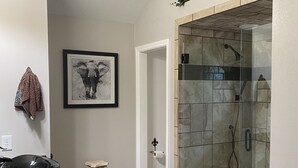 A bathroom with a glass-enclosed shower, a black countertop, a framed elephant artwork, and a door leading to another room. - Brick home in a quiet quaint neighborhood. Just minutes off the beach express. (Foley)