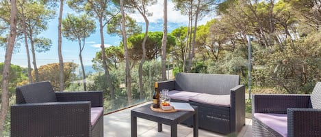 An outdoor seating area with wicker furniture and a table, overlooking a scenic landscape with trees and a view of the sea.
