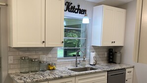 A modern kitchen with white cabinets, a granite countertop, a built-in dishwasher, and a window with a view of greenery. - 🌴Tropical Retreat in Sarasota  (Sarasota)