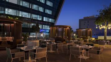 Un área de comedor al aire libre con mesas y sillas, una terraza de madera y un edificio moderno al fondo.