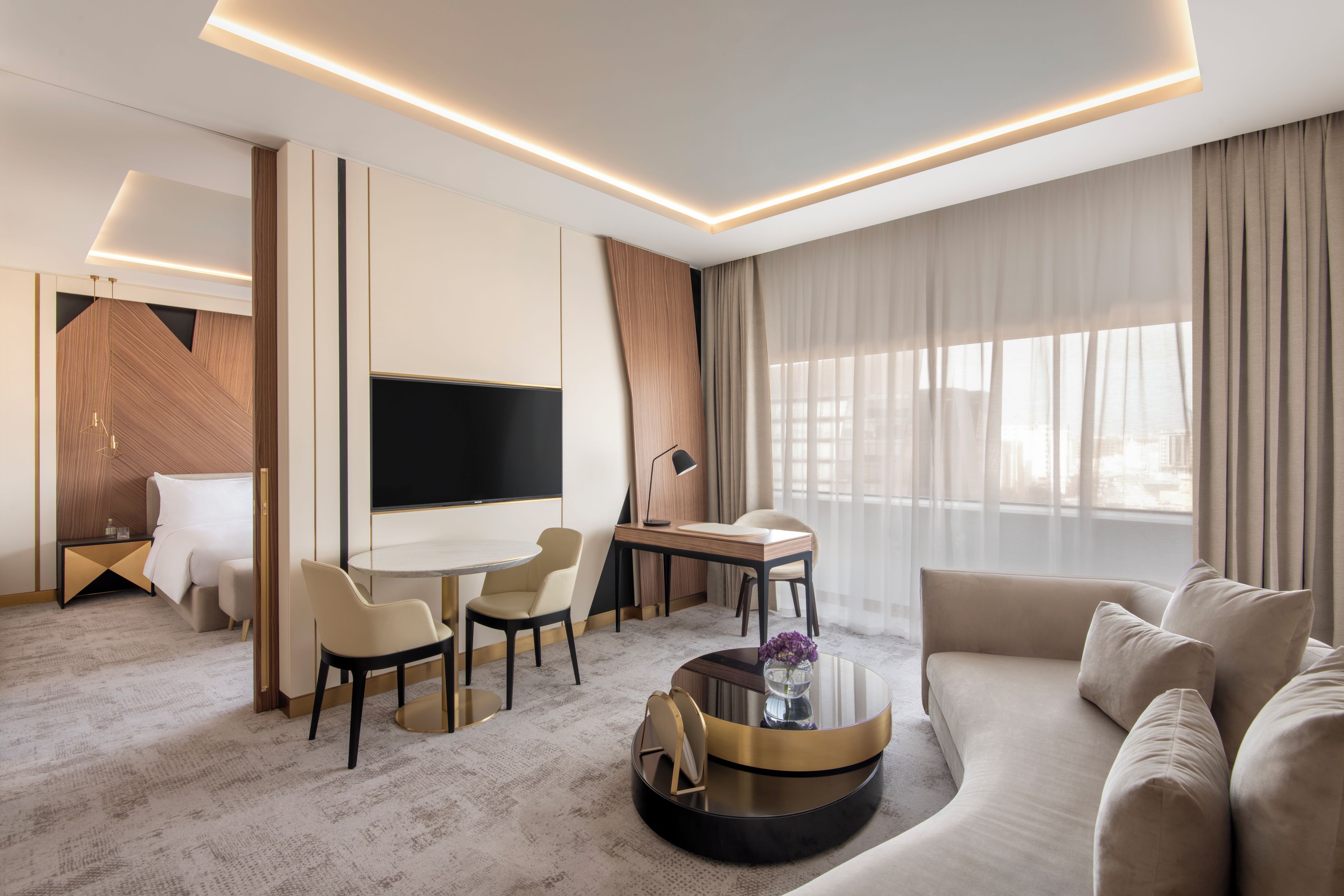 Una habitación de hotel moderna con televisión de pantalla plana, un sofá, un comedor con sillas, una mesa de centro redonda y un ventanal con cortinas.