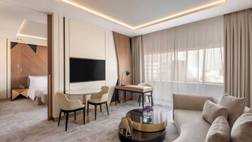 Una habitación de hotel moderna con televisión de pantalla plana, un sofá, un comedor con sillas, una mesa de centro redonda y un ventanal con cortinas.