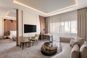 Una habitación de hotel moderna con televisión de pantalla plana, un sofá, un comedor con sillas, una mesa de centro redonda y un ventanal con cortinas.