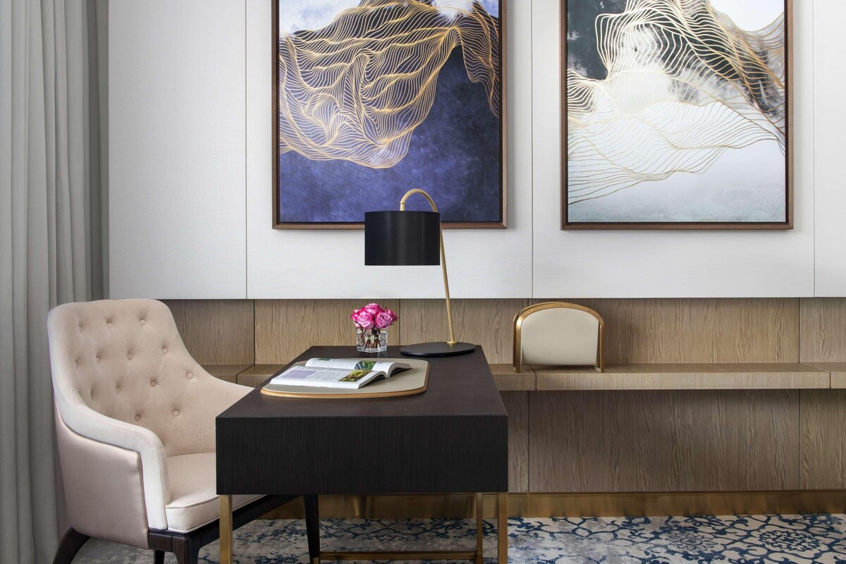 Una habitación moderna con un sillón capitoné, un escritorio oscuro con una lámpara y dos cuadros abstractos en la pared.
