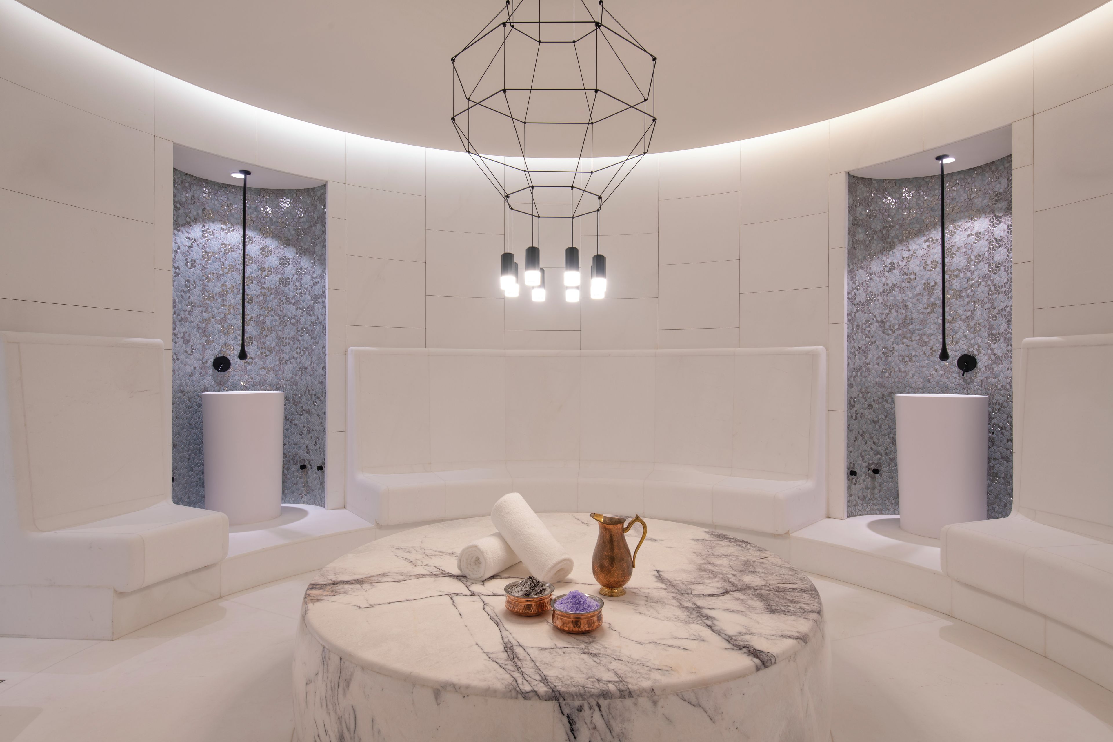A modern spa room with a circular marble table, two white towels, a copper pitcher, and a small bowl of bath salts.