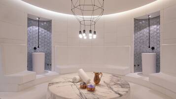 A modern spa room with a circular marble table, two white towels, a copper pitcher, and a small bowl of bath salts.