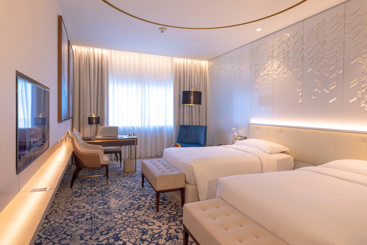 Habitación de hotel con dos camas, un escritorio y silla, una mesita y un amplio ventanal con cortinas.