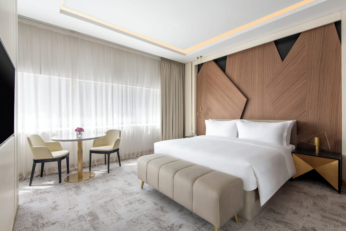 Habitación de hotel con una cama grande, una mesa redonda con sillas y vistas de la ciudad a través de amplias ventanas.