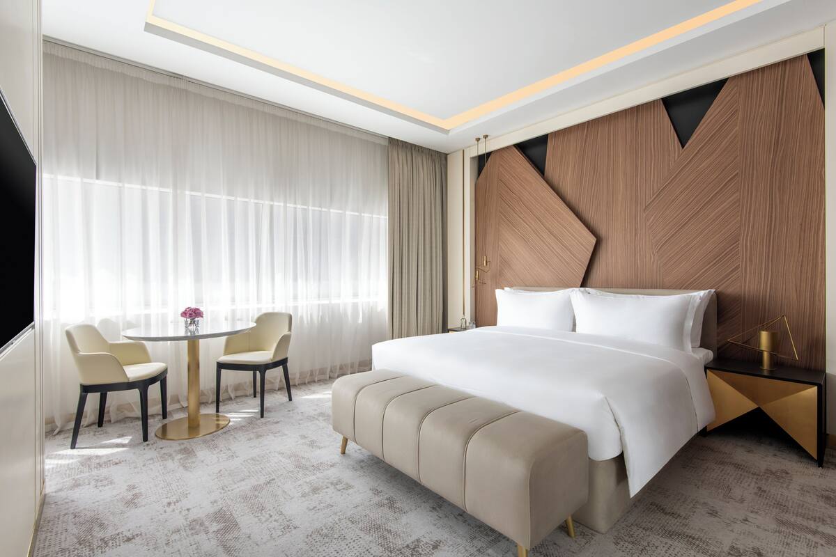 A hotel room with a large bed, a round table with chairs, a TV, and a view of the city.
