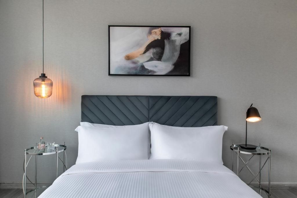 Una habitación de hotel moderna con una cama grande, un cabecero, un cuadro abstracto enmarcado, una mesa redonda con una lámpara y una lámpara colgante.