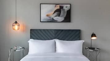 Una habitación de hotel moderna con una cama grande, un cabecero, un cuadro abstracto enmarcado, una mesa redonda con una lámpara y una lámpara colgante.