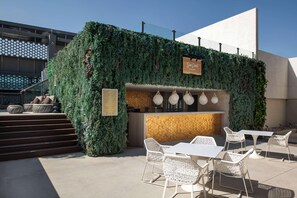 An outdoor seating area with white wicker chairs and a wooden counter, surrounded by lush green foliage. - Steigenberger Hotel Doha (Doha)