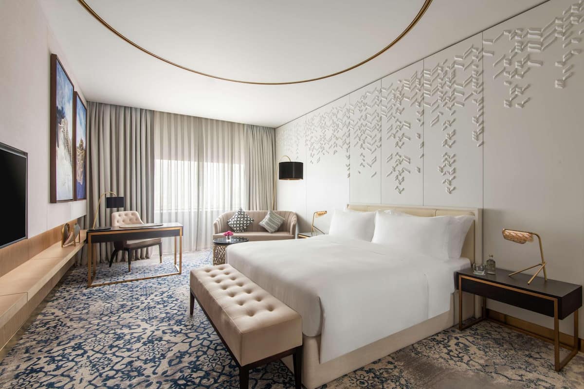 Habitación de hotel con una cama grande, un escritorio, una silla, un banco, un televisor y una alfombra estampada.