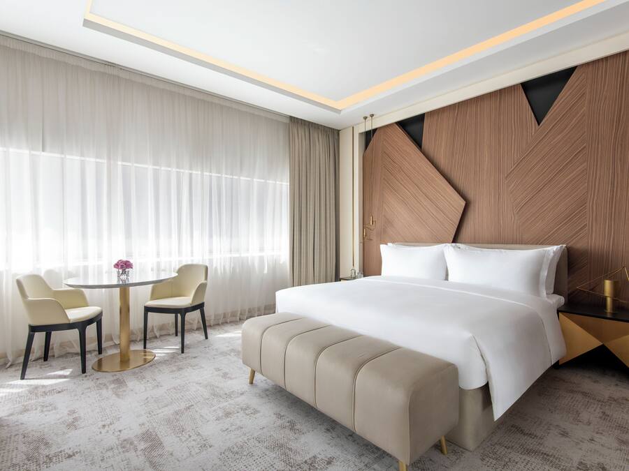 A hotel room with a large bed, a round table with chairs, and a view of the city through large windows.