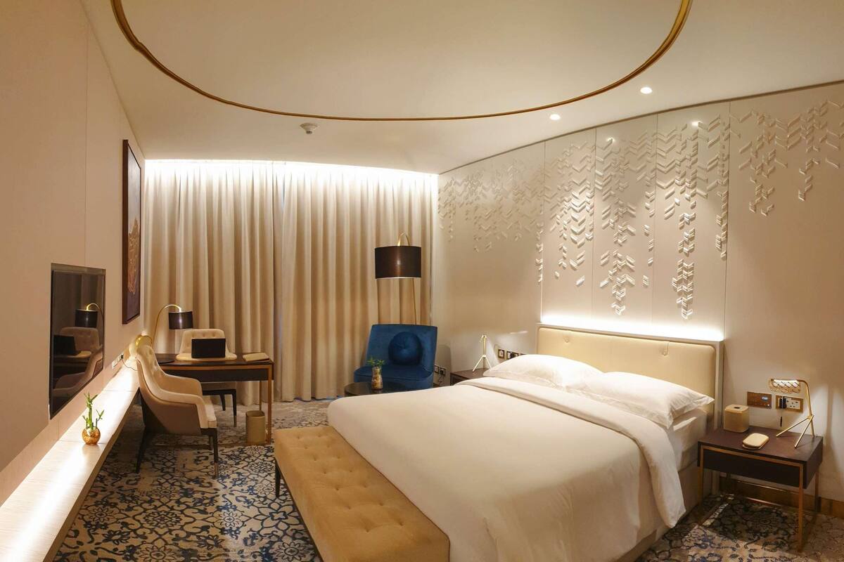 A hotel room with a large bed, a desk with a computer, a chair, a small table, and a decorative wall panel.
