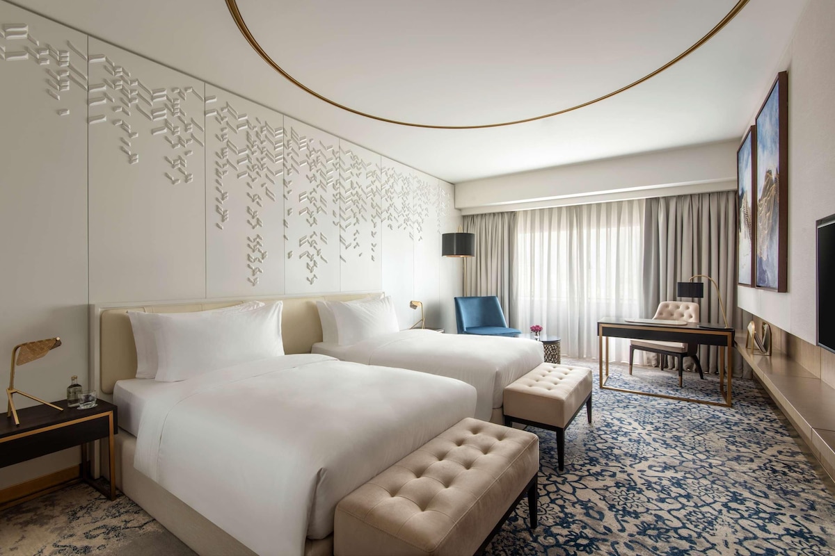 Habitación de hotel con dos camas, un escritorio, una silla, una mesita y alfombra estampada.