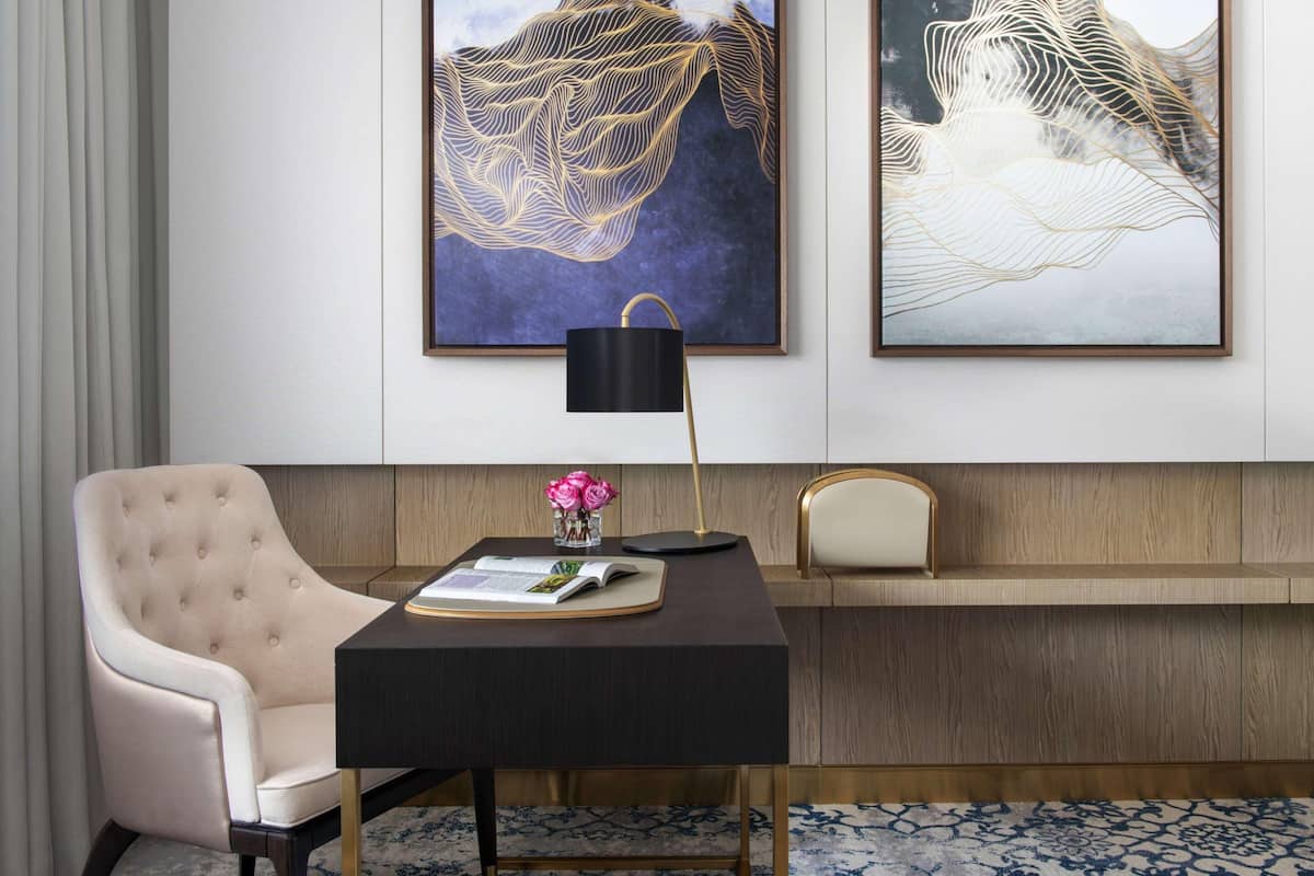 Una habitación moderna con un sillón capitoné, un escritorio oscuro con una lámpara y dos cuadros abstractos en la pared.